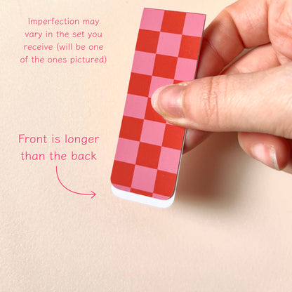 A hand holds a magnetic bookmark with a pink and red checkerboard pattern against a light beige background, with pink text noting that imperfections may vary and the front is longer than the back.