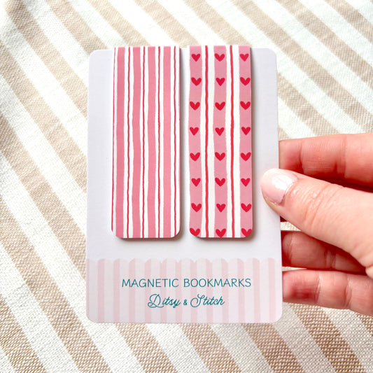 Magnetic bookmarks with pink stripes and hearts held by a hand on a striped fabric background