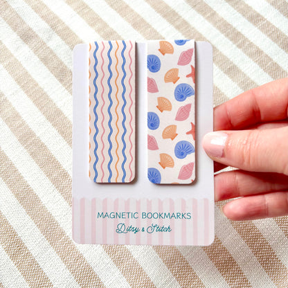 Two magnetic bookmarks with shell pattern design held by a hand on a striped background