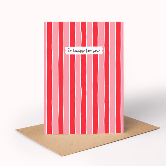 A greeting card with vertical pink and red stripes and a small white label reading 'So happy for you!' in black script, shown standing upright with a brown kraft envelope on a white background.
