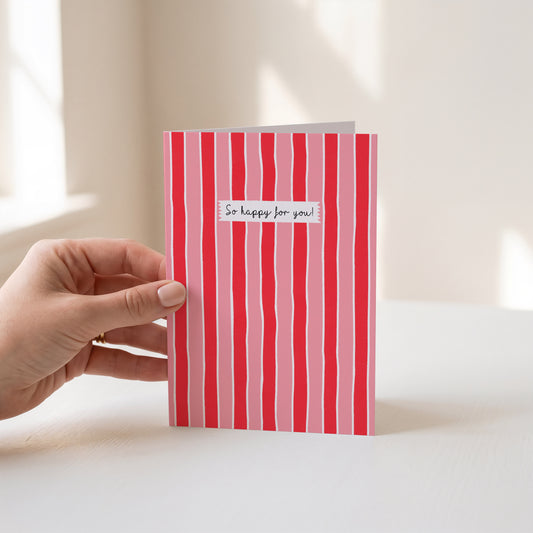A manicured hand holds a greeting card with vertical pink and red stripes and a small white label reading 'So happy for you!' in black script, against a soft light background.