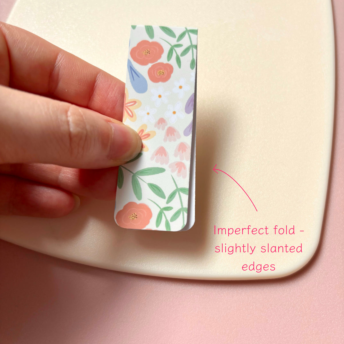 A hand holds magnetic bookmark with a pastel floral print against a pink background, with a cream surface behind it and pink text noting imperfect fold slightly slanted edges.