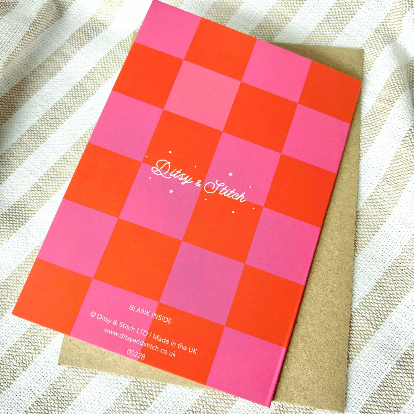 Checkered card with pink and orange squares and 'Ditsy & Stitch' branding on the back on a textured fabric background.