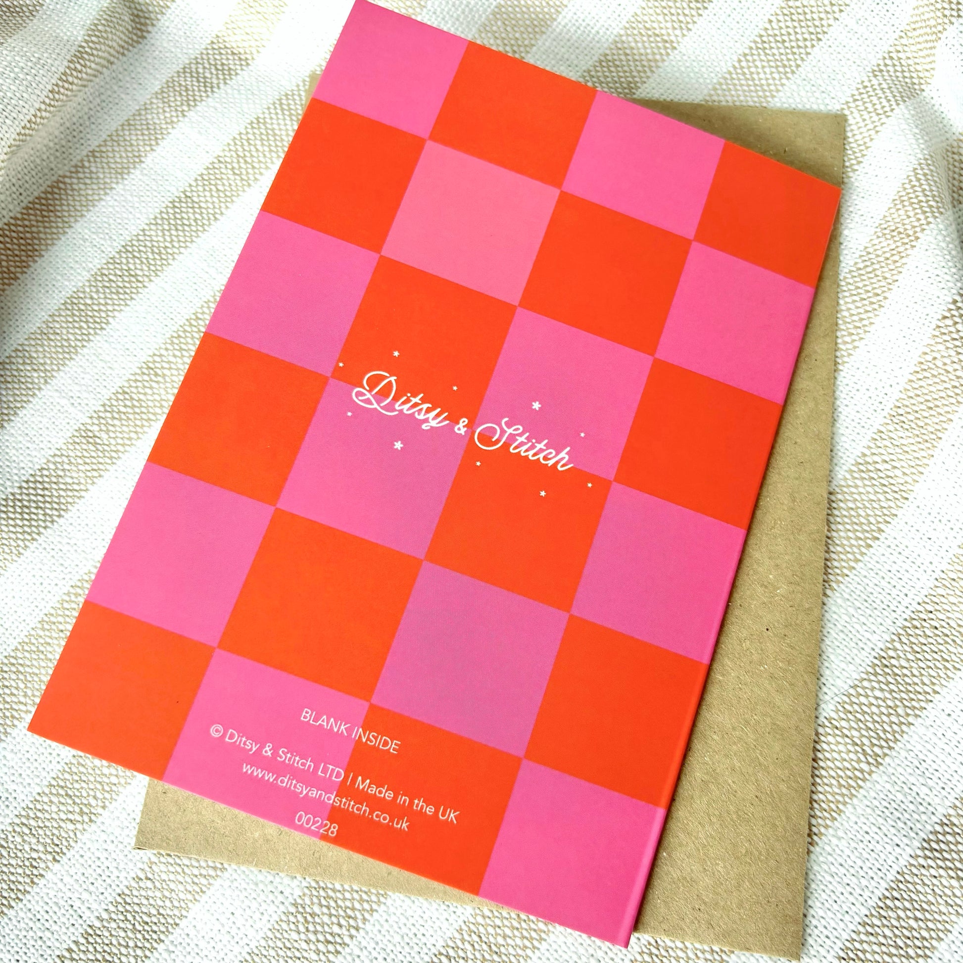 Checkered card with pink and orange squares and 'Ditsy & Stitch' branding on the back on a textured fabric background.