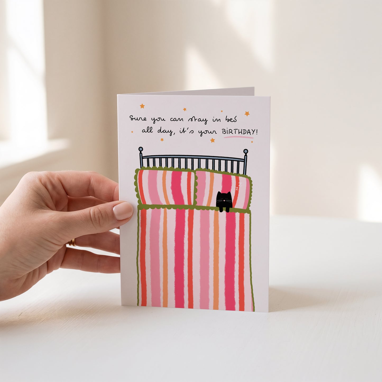 A hand holds a greeting card with a whimsical illustration of a bed with pink and orange striped bedding and a small black cat. The card has handwritten text that reads, 'Sure you can stay in bed all day, it's your BIRTHDAY!'.