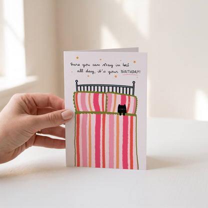 A hand holds a greeting card with a whimsical illustration of a bed with pink and orange striped bedding and a small black cat. The card has handwritten text that reads, 'Sure you can stay in bed all day, it's your BIRTHDAY!'.