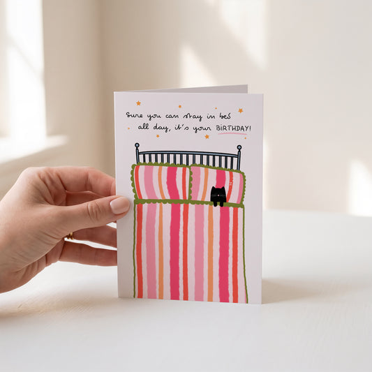 A hand holds a greeting card with a whimsical illustration of a bed with pink and orange striped bedding and a small black cat. The card has handwritten text that reads, 'Sure you can stay in bed all day, it's your BIRTHDAY!'.