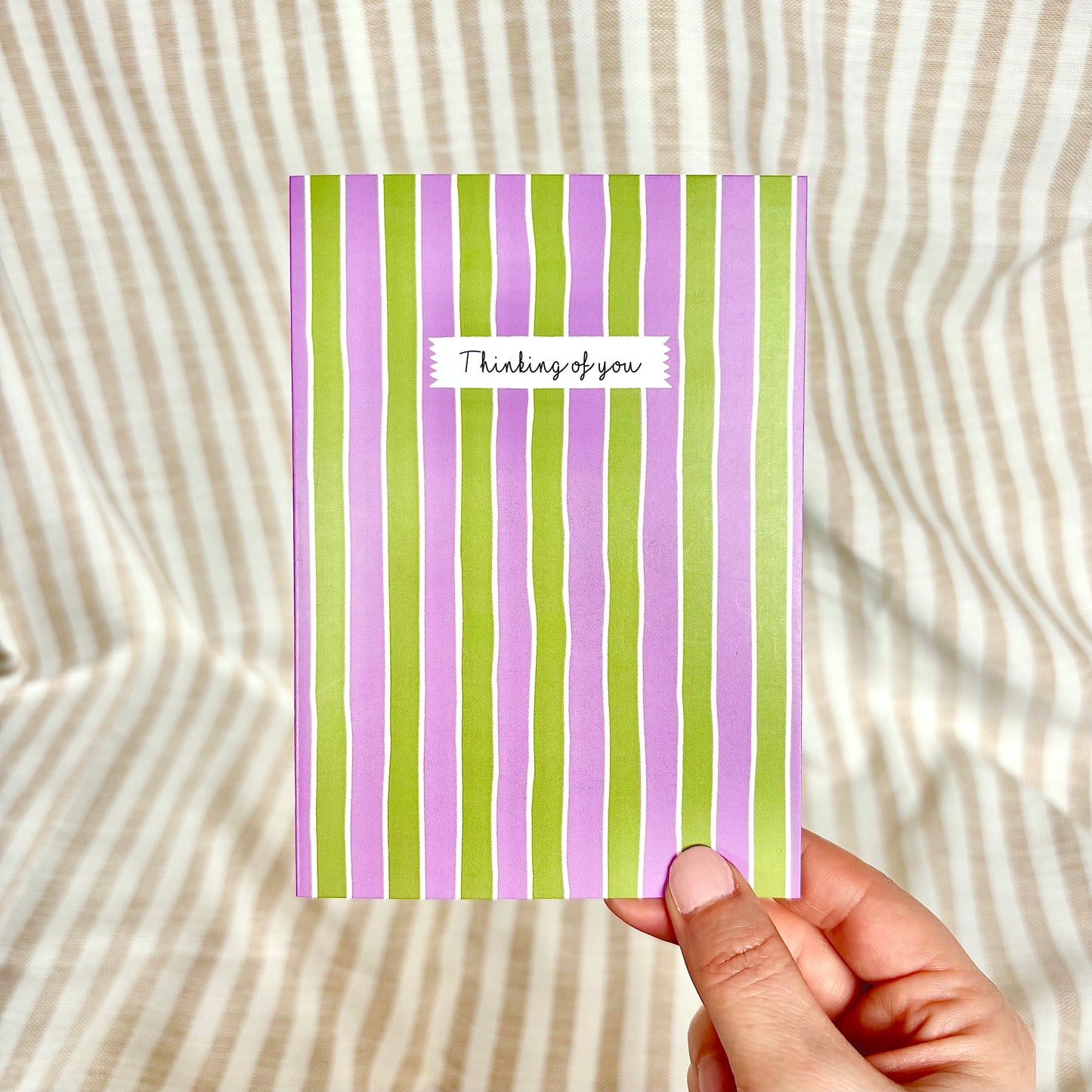 Hand holding a card with purple and green stripes and 'Thinking of you' text on a striped background.