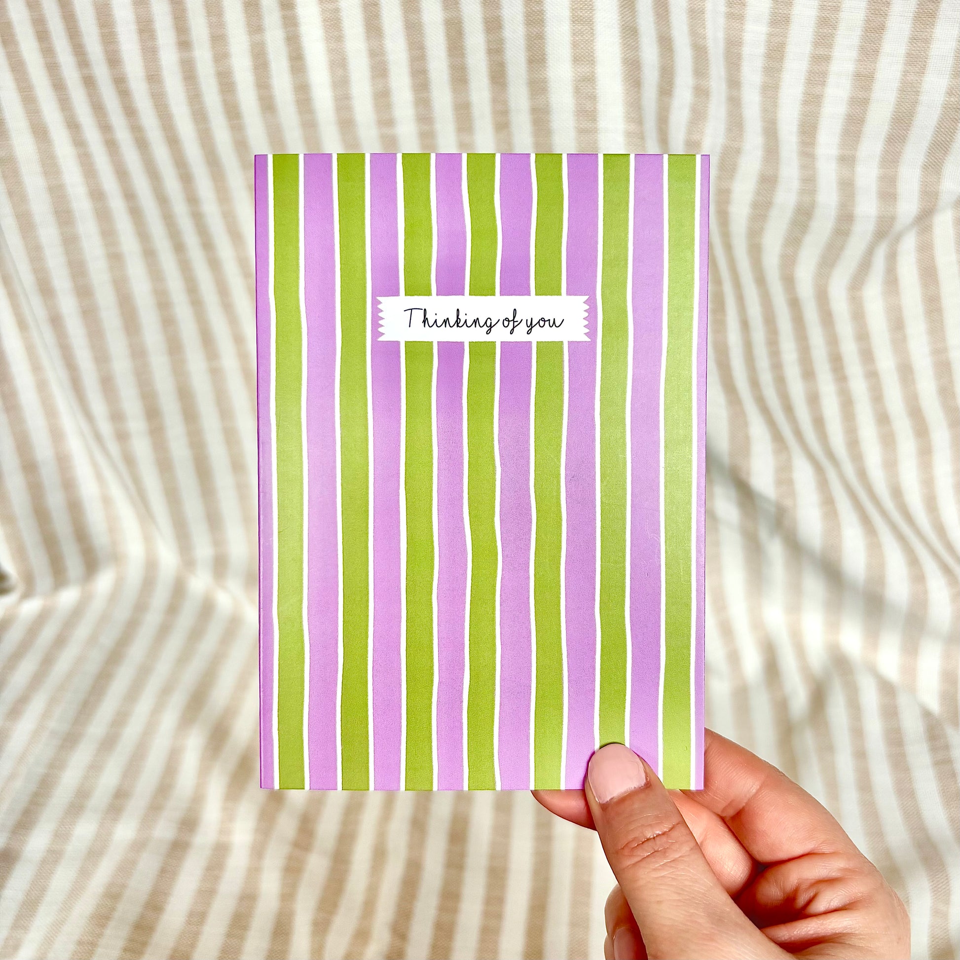 Hand holding a card with purple and green stripes and 'Thinking of you' text on a striped background.
