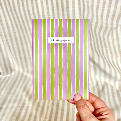 Hand holding a card with purple and green stripes and 'Thinking of you' text on a striped background.