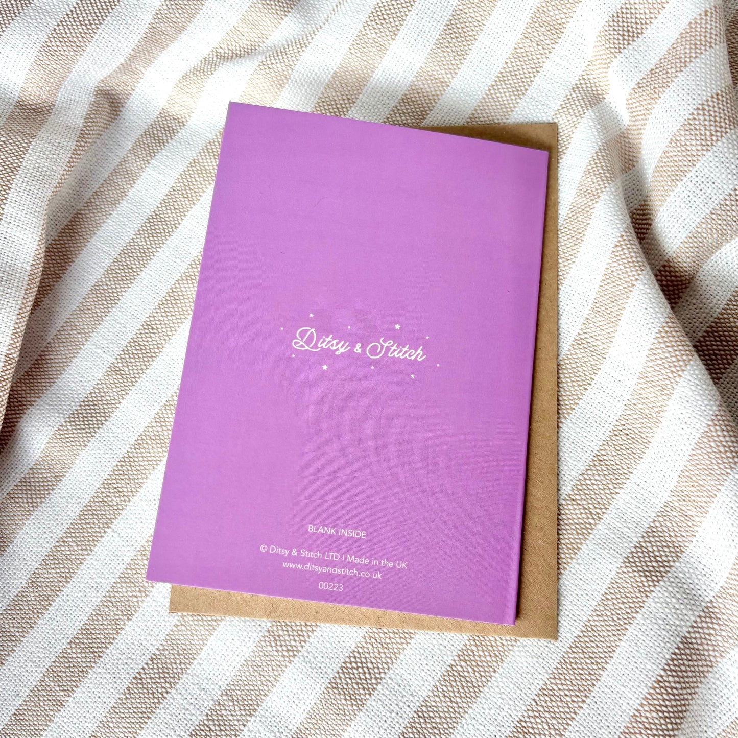 Purple greeting card with 'Daisy & Stitch' branding on the back, on a striped fabric background