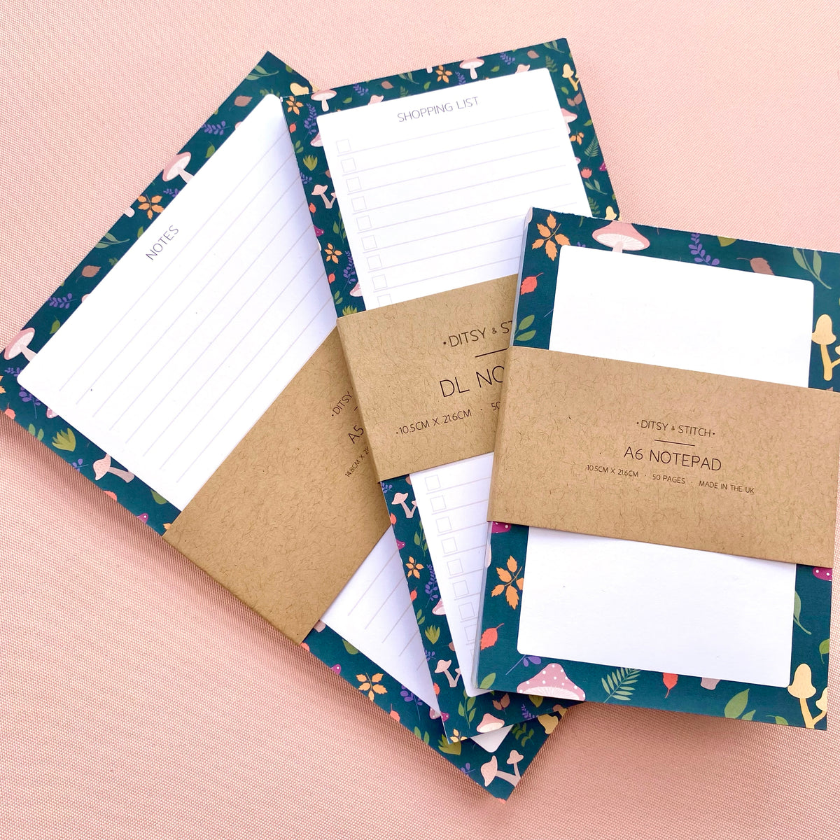 Three autumn notepads with kraft paper bands are arranged on a soft pink background. The notepads have dark teal borders with leaves and toadstools and white writing pages.