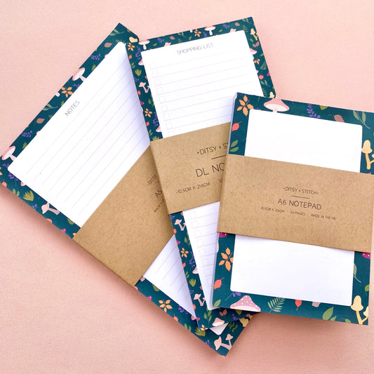 Three autumn notepads with kraft paper bands are arranged on a soft pink background. The notepads have dark teal borders with leaves and toadstools and white writing pages.