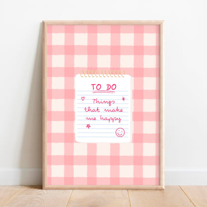 A framed wall art print with a pink and white gingham pattern background, featuring a white notepad graphic in the centre with a handwritten-style 'TO DO' list with ‘Things that make me happy’ and smiley face. The frame is leaning against a white wall on a light wood floor.