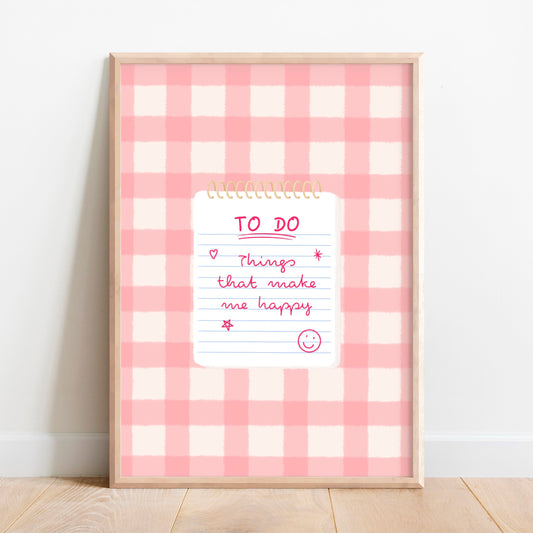 A framed wall art print with a pink and white gingham pattern background, featuring a white notepad graphic in the centre with a handwritten-style 'TO DO' list with ‘Things that make me happy’ and smiley face. The frame is leaning against a white wall on a light wood floor.