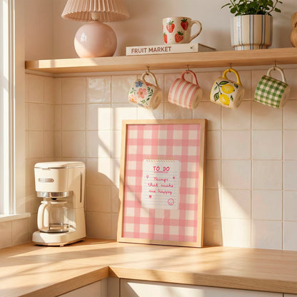 A cosy kitchen corner with a pink gingham framed sign on a wooden counter, hanging floral mugs from hooks on a tiled wall, a cream coffee maker, and a shelf with mugs and a plant in the background.
