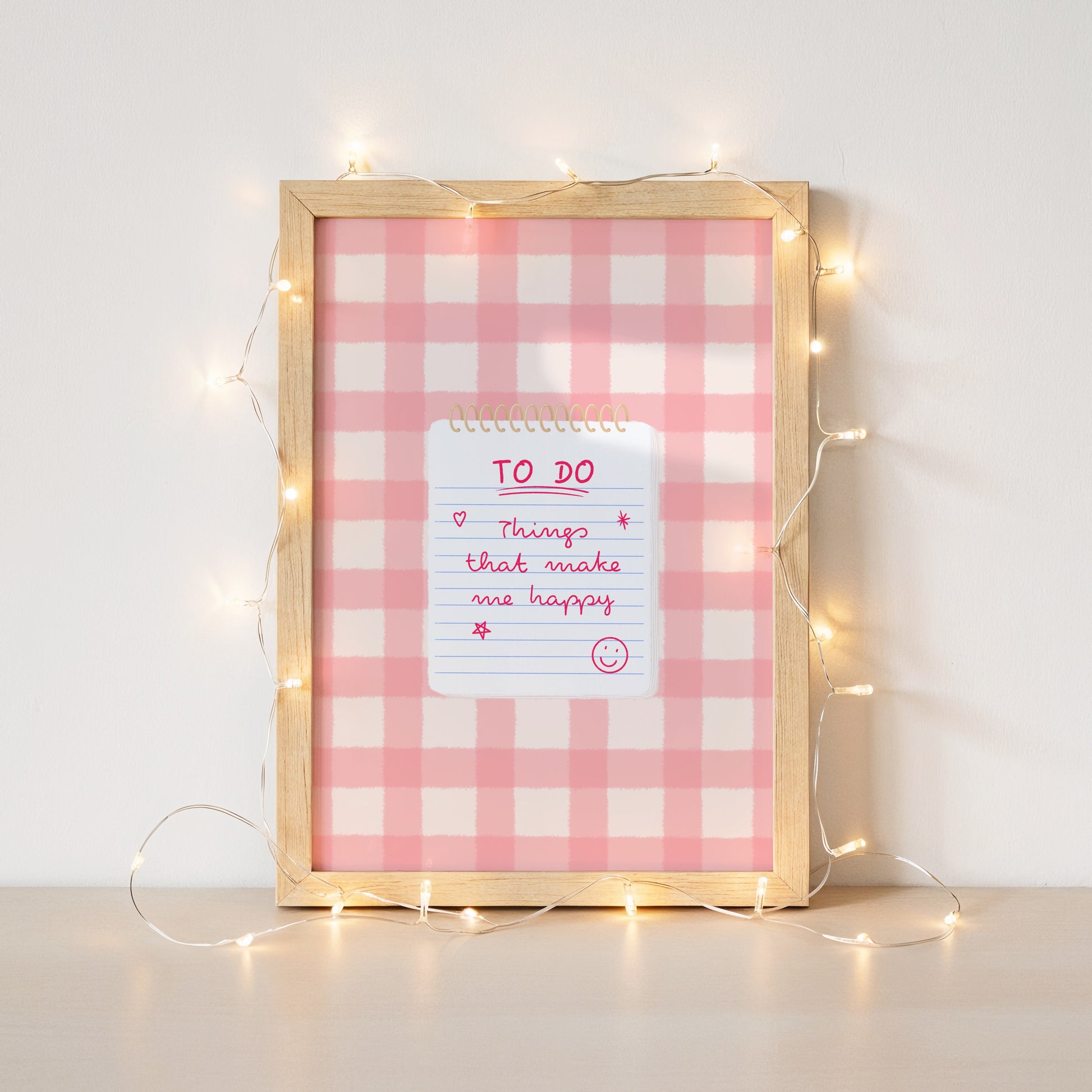 A square framed wall art piece with a pink and white gingham pattern, a light wood frame, and a small white notepad reading 'TO DO Things that make me happy' with pink handwritten text and stars. Warm string lights are draped around the frame against a white wall.