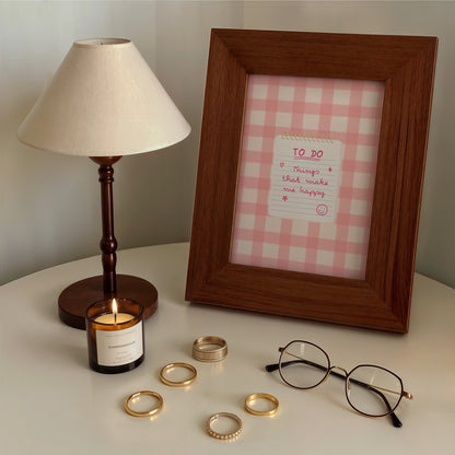 A wooden picture frame with a pink gingham check pattern and a handwritten 'TO DO' note, styled on a white table next to a lamp, candle, rings, and eyeglasses.