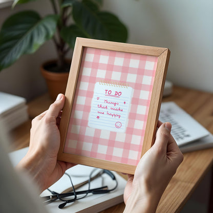 A person holds a light wood framed notepad with a pink and white gingham pattern and a handwritten 'TO DO' note. The scene is set on a wooden desk with a potted plant, glasses, and books in the background.