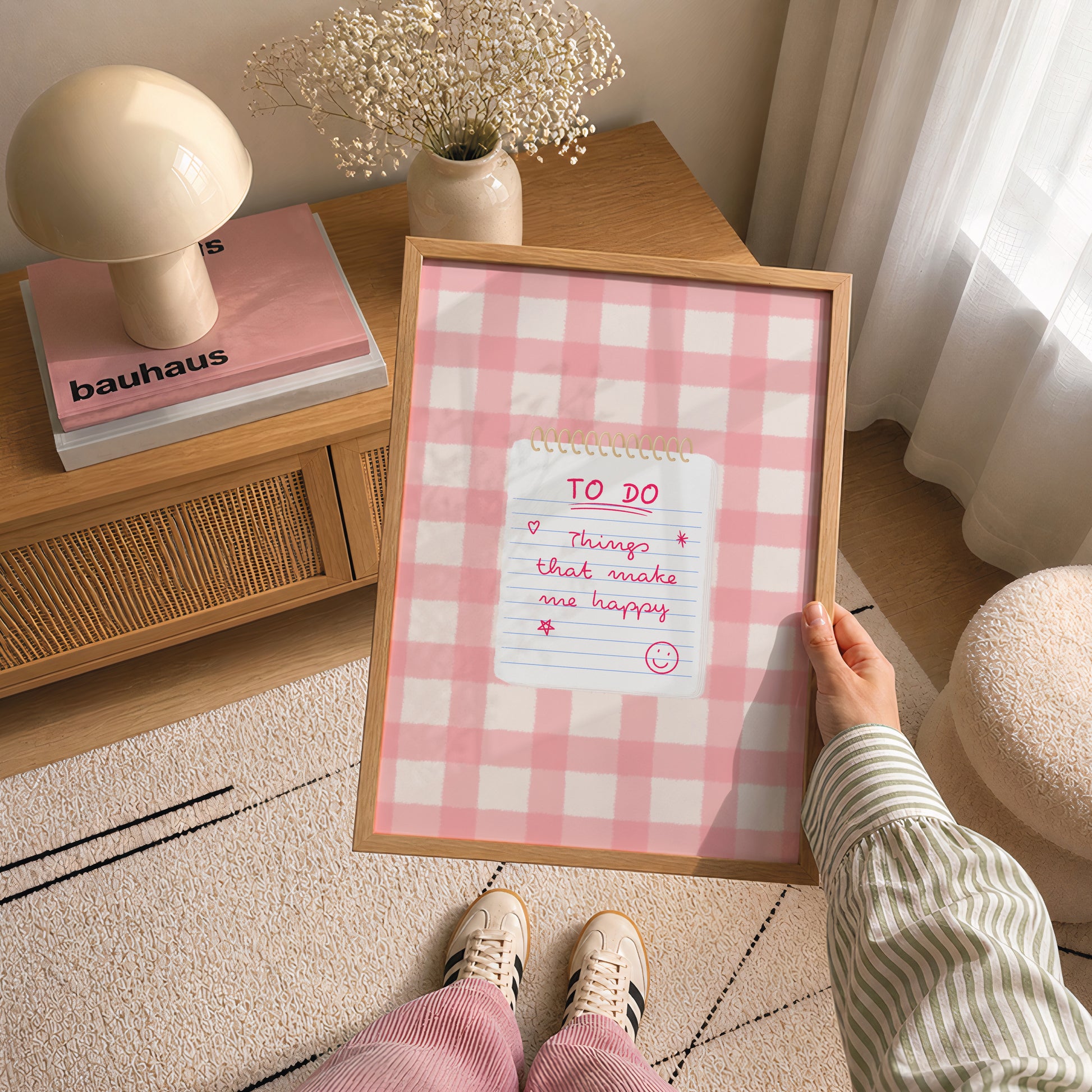 A person holds a framed art print featuring a happy to do list on a pink gingham background. The scene is set in a cozy, modern room with a wooden side table, a mushroom-shaped lamp, and a patterned rug.