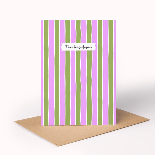 A vertical greeting card with alternating pink and green wavy vertical stripes, featuring a small white label in the center that reads 'Thank you' in black script. A plain brown envelope lies flat on a white surface behind the card.