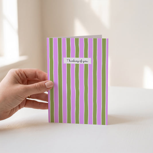 A hand holds a greeting card with vertical pink and olive green stripes. A small white label on the card reads 'Thinking of you'.