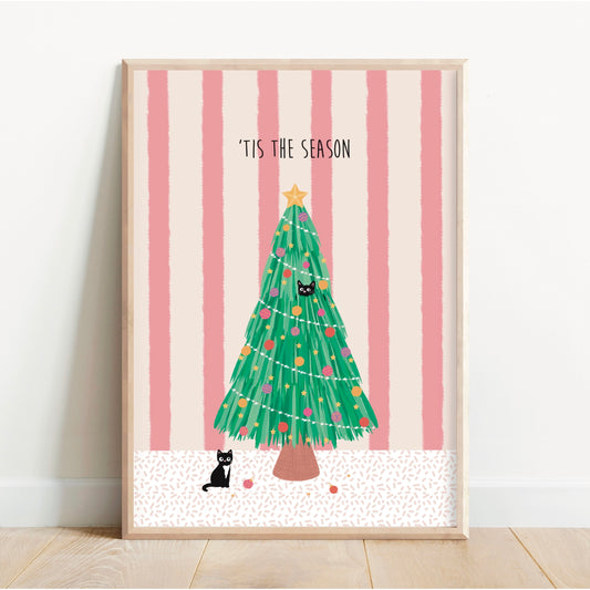 A framed art print featuring a green Christmas tree with colourful ornaments against pink and white vertical stripes, with the text "TIS THE SEASON" at the top. The print is displayed leaning against a white wall on a light wood floor.