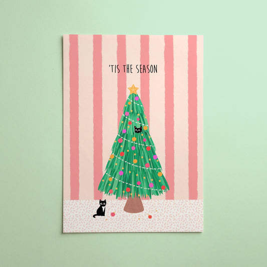 A vertical art print featuring a hand-drawn green Christmas tree with colourful ornaments, a gold star topper, and two black cats, with the text 'TIS THE SEASON' at the top. The card is set against a background of pink vertical stripes and is photographed against a solid mint green background.