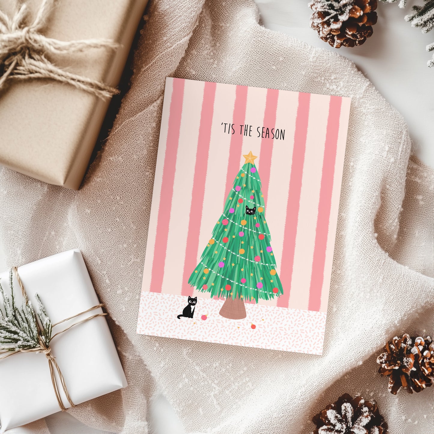 Christmas card with a tree and cat design on a striped background, surrounded by wrapped gifts and pine cones.