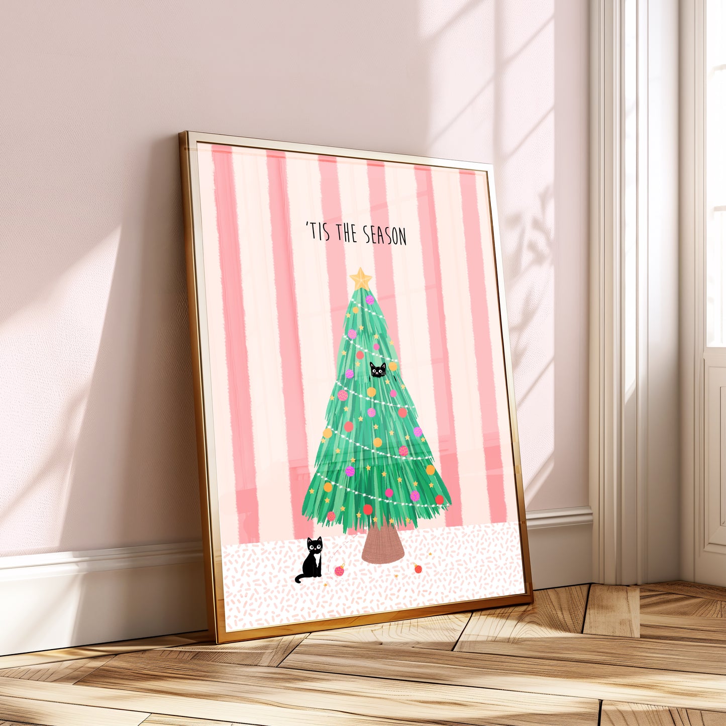 Framed artwork with a Christmas tree, black cats and text on a pink striped background, placed on a wooden floor.