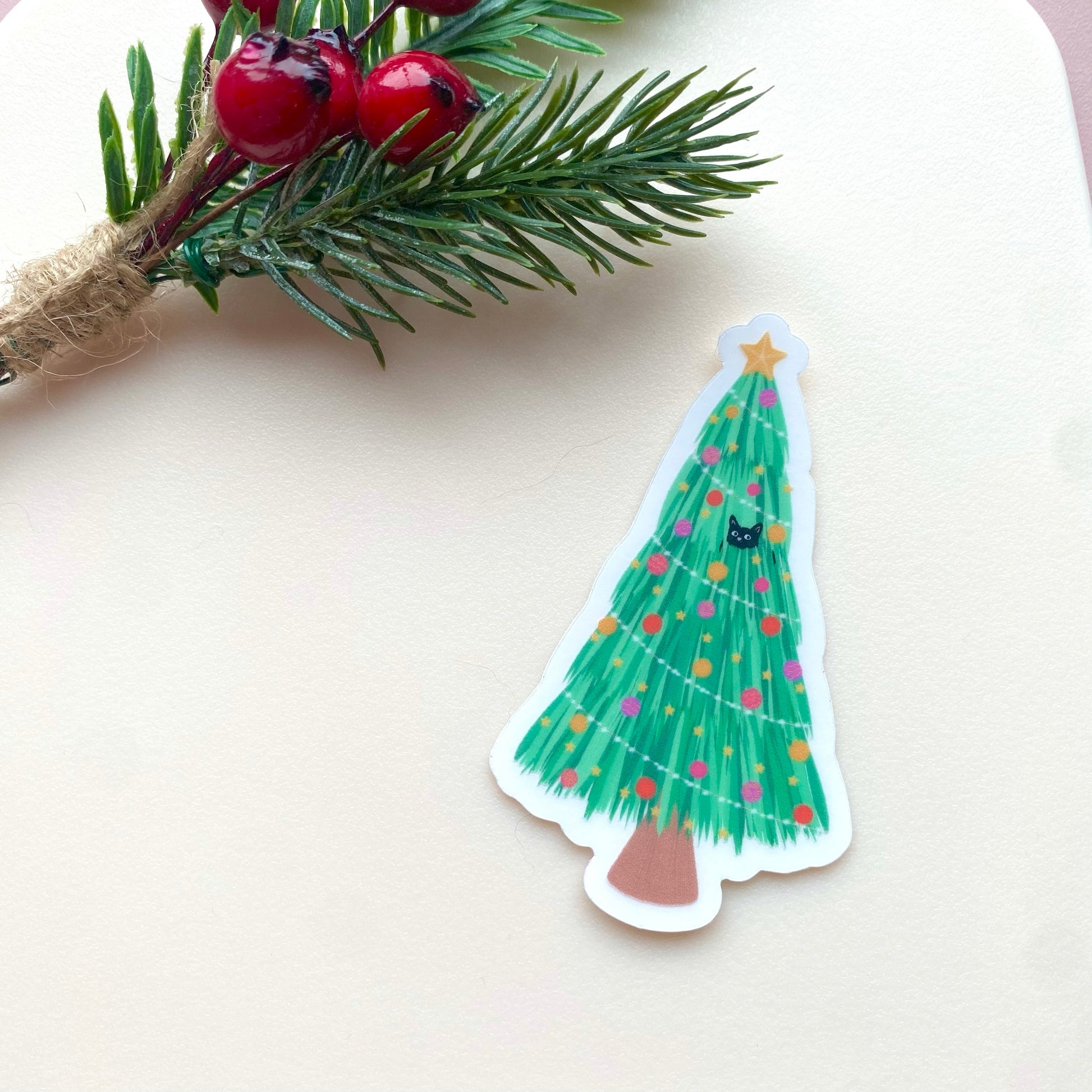 Sticker of a Christmas tree with colourful decorations and a cat in the branches with another under the tree. On a white background with a branch and berries.