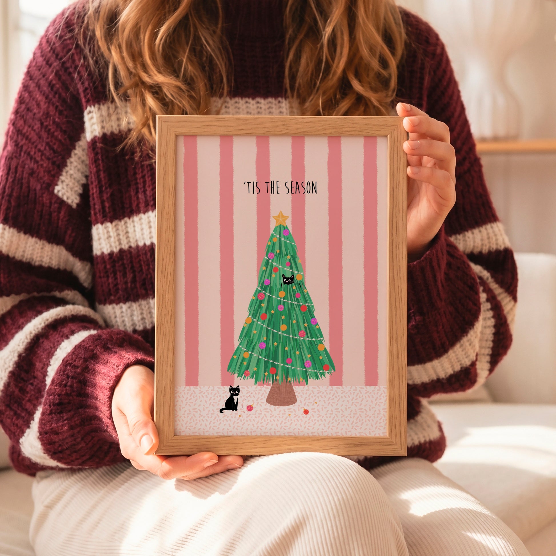 A person in a maroon and cream striped knit sweater holds a light wood framed art print featuring a green Christmas tree with ornaments, two black cats, pink vertical stripes, and the text 'IT'S THE SEASON'.