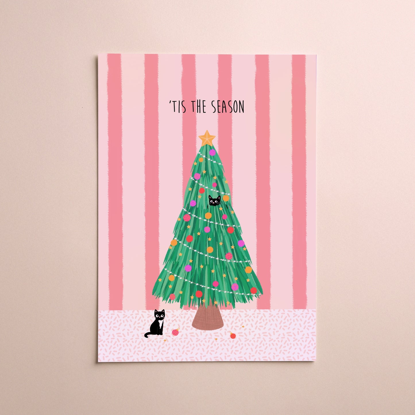 A vertical greeting card featuring a festive Christmas tree illustration with the text '“TIS THE SEASON' at the top, displayed against a soft pink striped background with two small black cats on the card and a beige wall behind it.