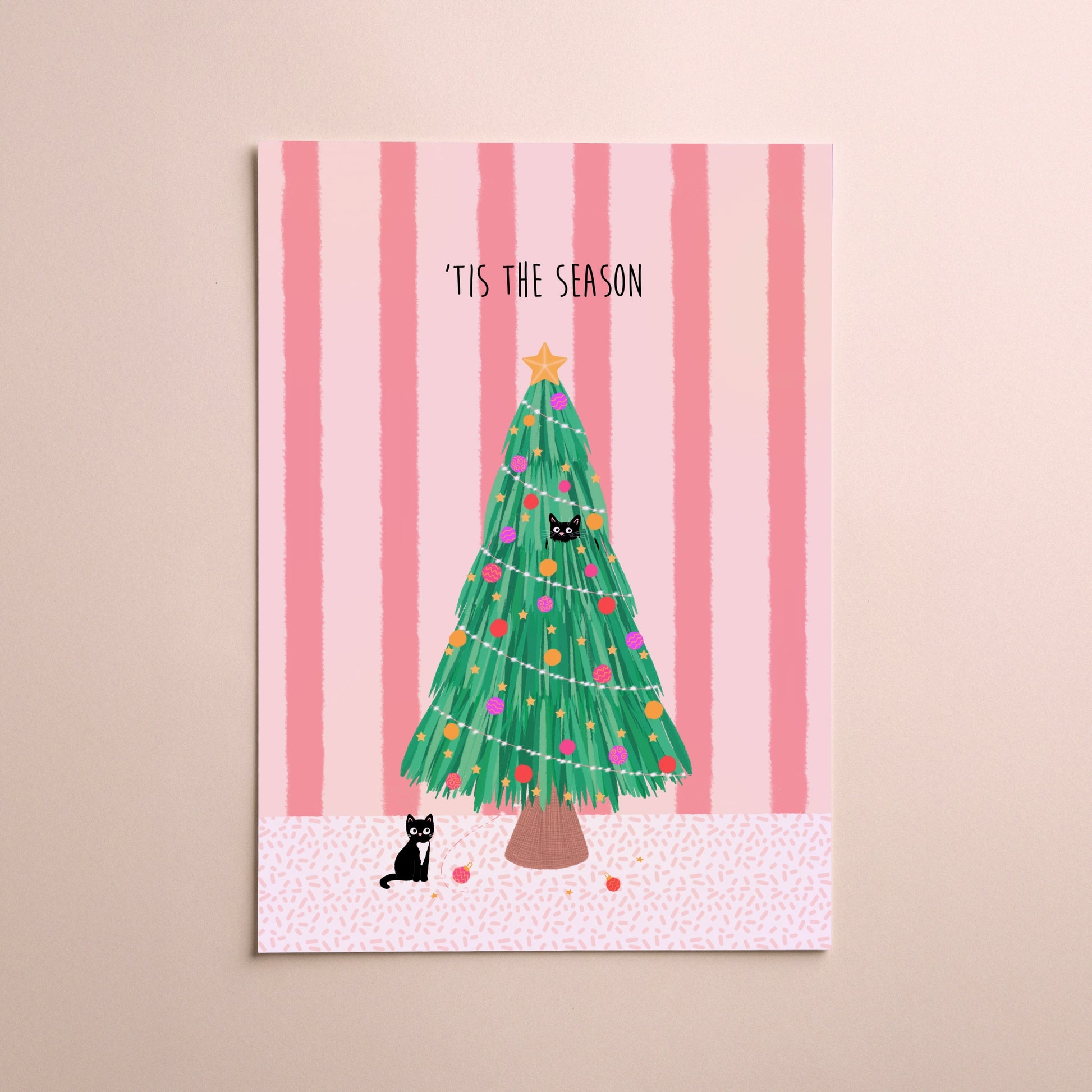 A vertical greeting card featuring a festive Christmas tree illustration with the text '“TIS THE SEASON' at the top, displayed against a soft pink striped background with two small black cats on the card and a beige wall behind it.