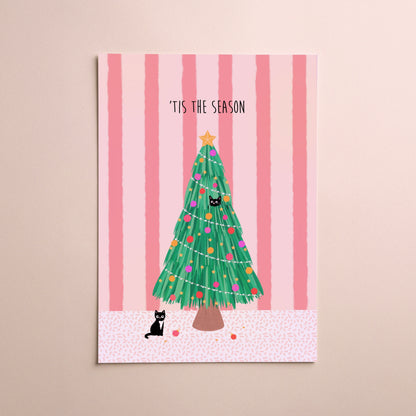 A vertical greeting card featuring a festive Christmas tree illustration with the text '“TIS THE SEASON' at the top, displayed against a soft pink striped background with two small black cats on the card and a beige wall behind it.