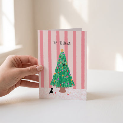 A hand holds a Christmas greeting card featuring a green Christmas tree with colourful ornaments against a pink and white striped background, with the text 'IT'S THE SEASON'.