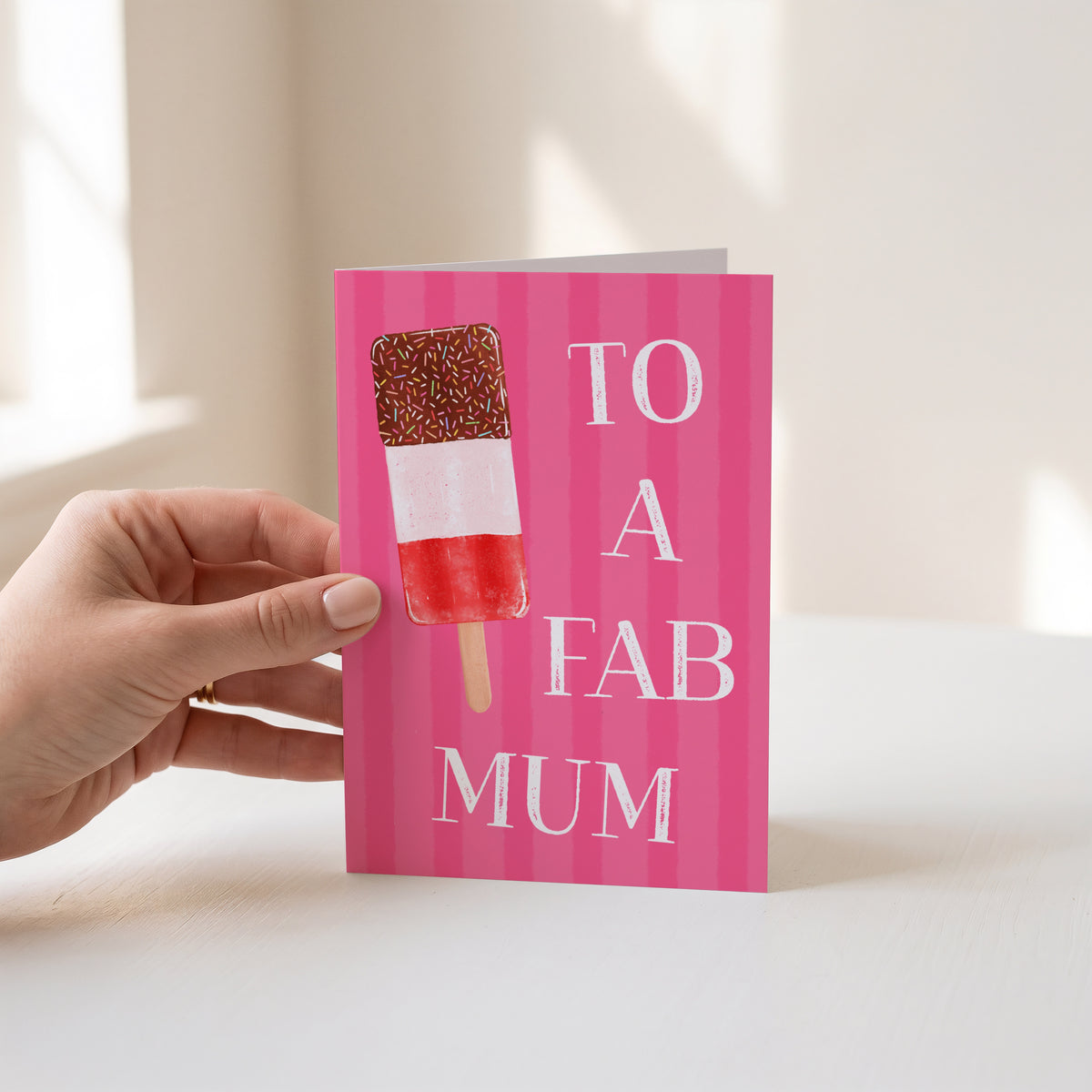 A hand holds a pink striped greeting card that says 'TO A FAB MUM' next to an ice lolly illustration, set against a bright, softly lit indoor background.