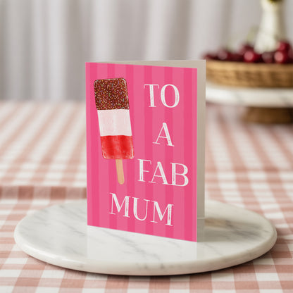 A pink greeting card with white text reading 'TO A FAB MUM' and an ice lolly illustration, standing on a round marble tray over a pink-and-white checkered tablecloth.