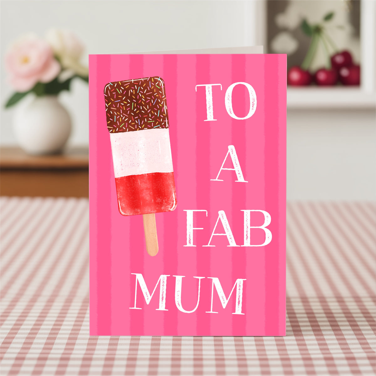A pink striped greeting card with a chocolate-dipped ice lolly illustration and the text 'TO A FAB MUM' in white, standing on a red-and-white checkered tablecloth with a blurred indoor background.