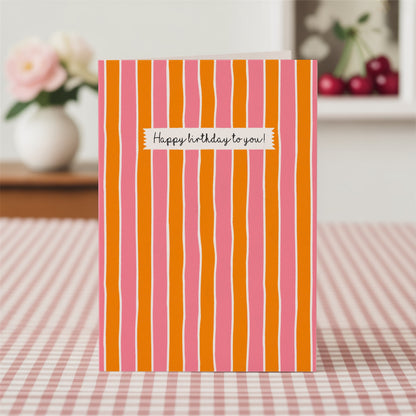 Striped birthday card with pink and orange vertical stripes on a checkered tablecloth.