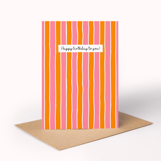 A vertical greeting card with wavy vertical stripes in pink, orange, and white, featuring a white label that reads 'Happy birthday to you!' in black script. The card is standing upright on a brown kraft envelope against a clean white background.