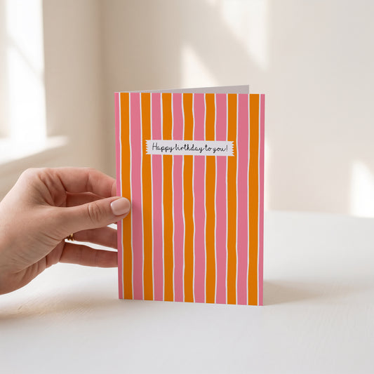 A hand holds a greeting card with vertical orange and pink stripes and a small white label reading 'Happy birthday to you!' against a softly lit indoor background.