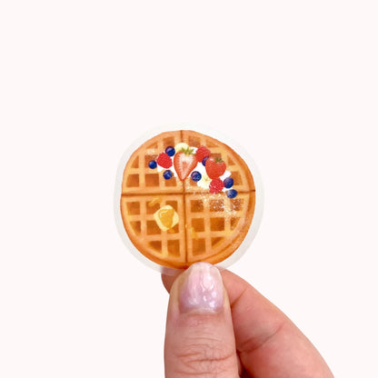 A transparent sticker with a detailed illustration of some breakfast waffles with syrup, butter and mixed summer berries