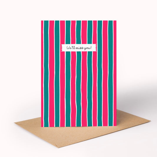 A greeting card with vertical pink and teal stripes and a white label reading 'We'll miss you!' in script, shown standing upright with a brown kraft envelope on a light gray background.