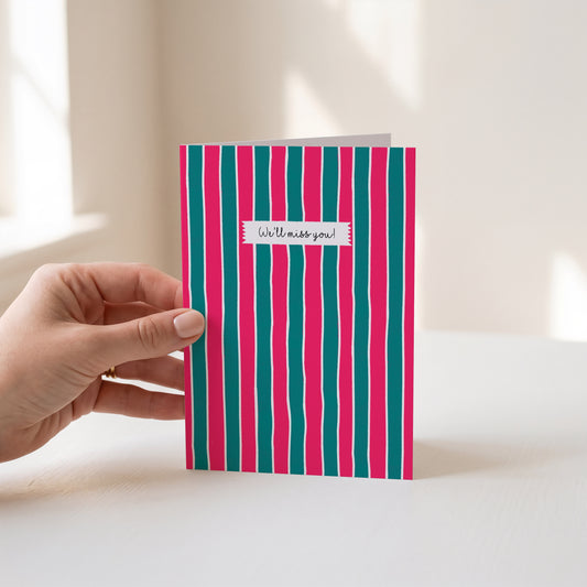 A hand holds a greeting card with vertical pink and teal stripes and a small white label reading 'We'll miss you!' against a softly lit indoor background.