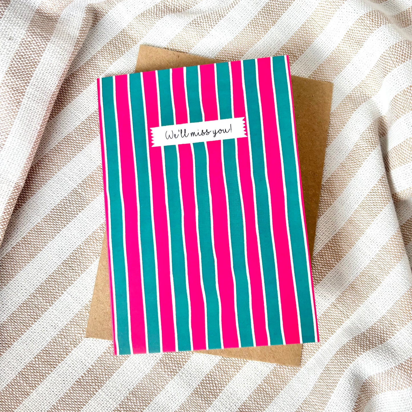 Striped card with 'We'll miss you!' message on a textured fabric background