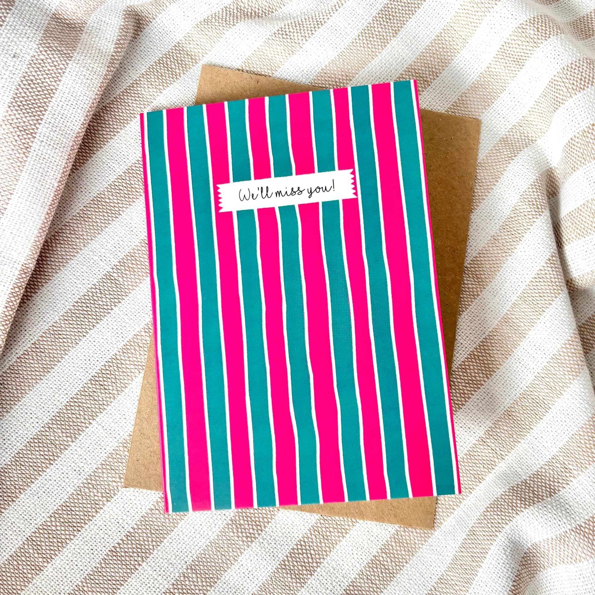 Striped card with 'We'll miss you!' message on a textured fabric background