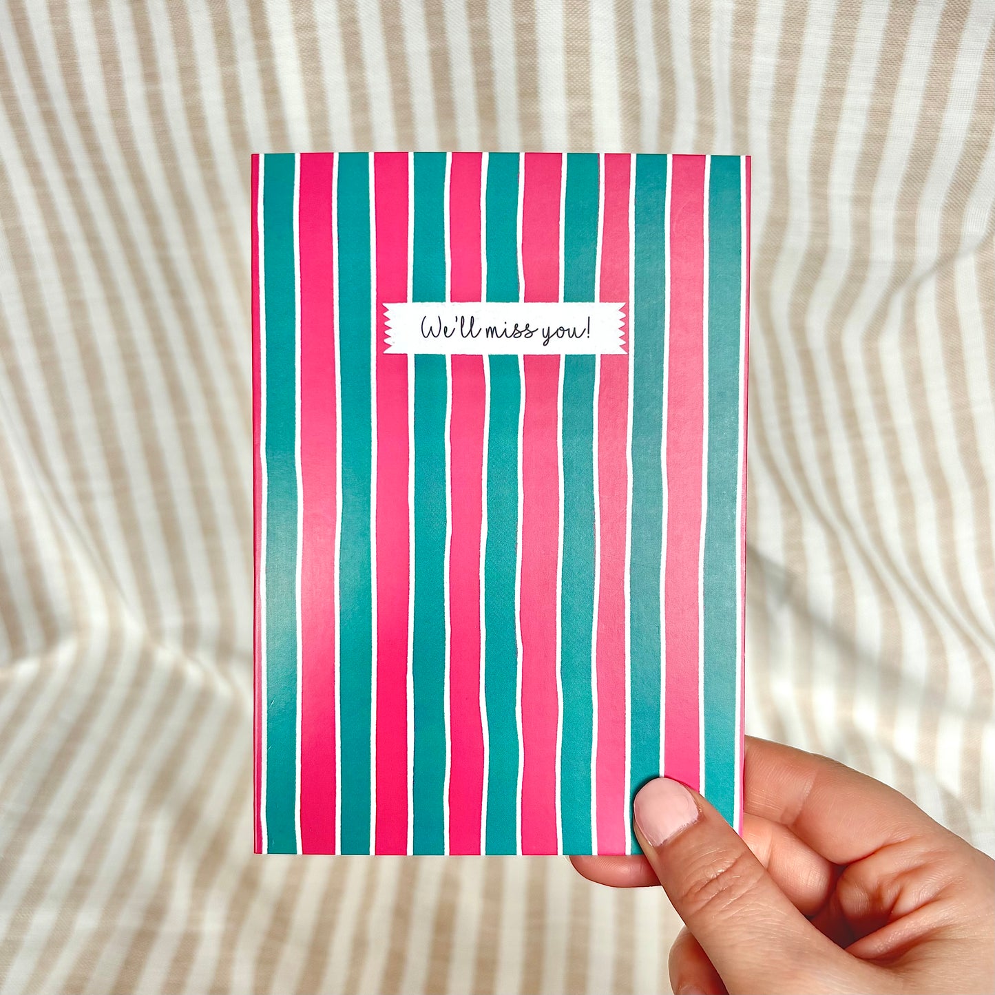 Striped card with 'We'll miss you!' message held by a hand on a textured background
