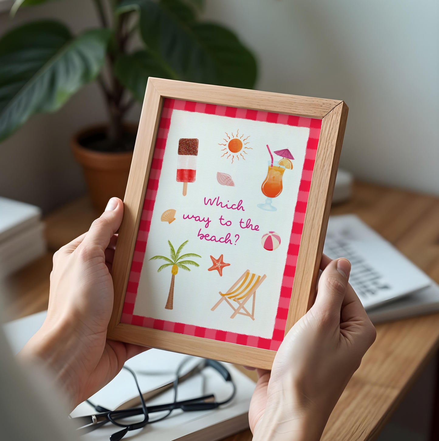 A person holds a framed summer-themed art print with illustrations of a beach umbrella, palm tree, deck chair, starfish, and cocktails, set against a cozy indoor desk background.
