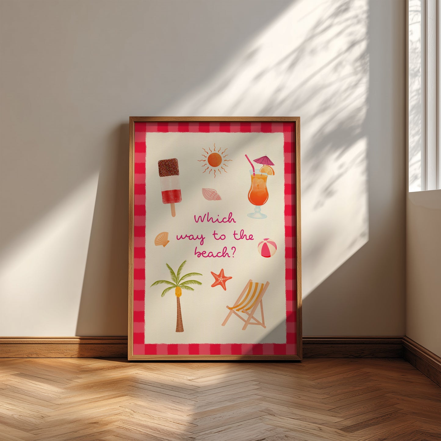 A framed beach-themed art print leaning against a wall on a wooden floor. The print features a pink and red striped border and colourful illustrations of a cocktail, palm tree, beach chair, starfish, and sun with the text 'Which way to the beach?'.